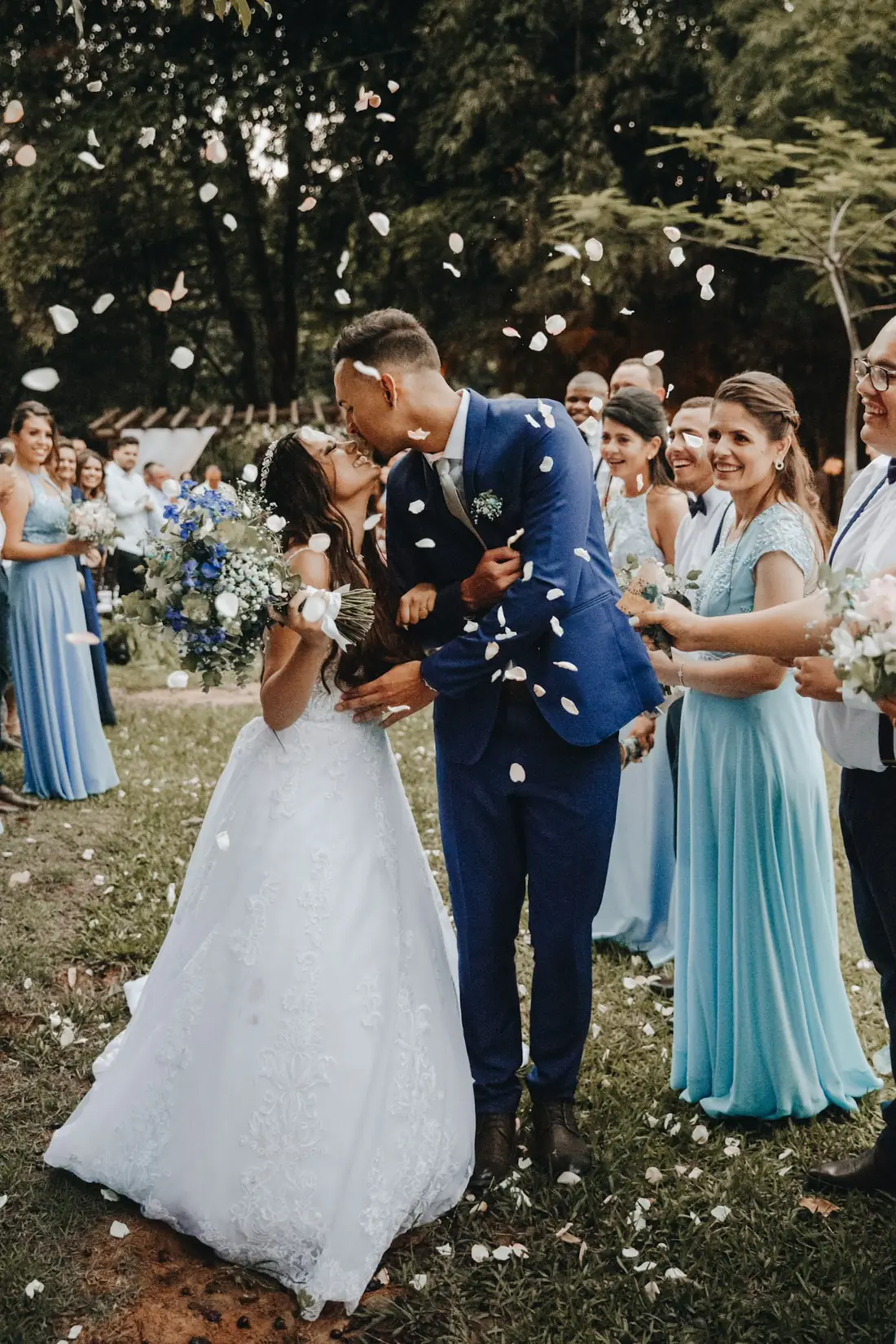 Novios besándose bajo una lluvia de pétalos rodeados de invitados